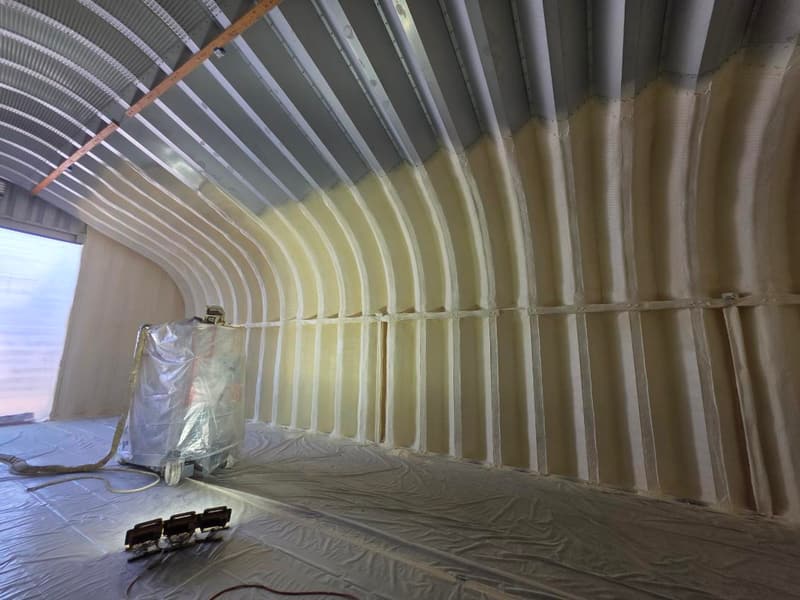 Arched metal building partway through spraying: foam on the lower curve, bare corrugated metal still showing overhead