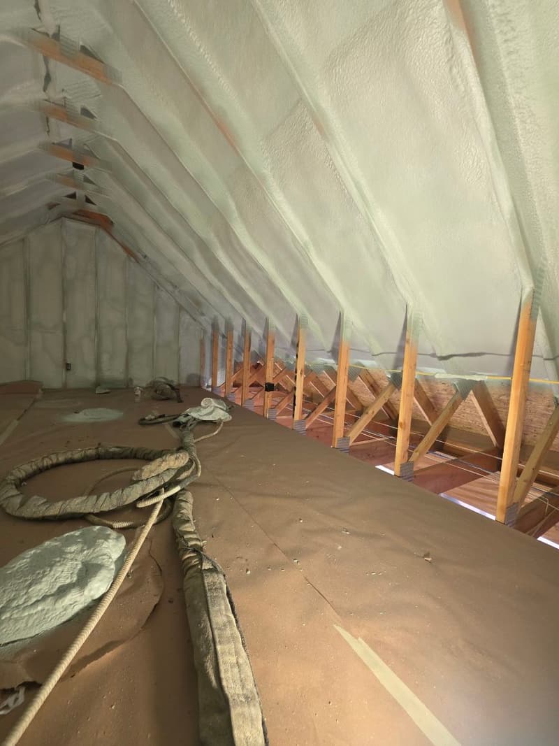 Attic with spray foam along the underside of the roof above the trusses and a coiled spray hose on the paper-covered floor