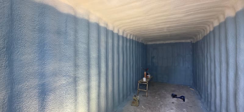 Long, narrow room with corrugated walls coated in blue-tinted spray foam and lighter foam across the ceiling