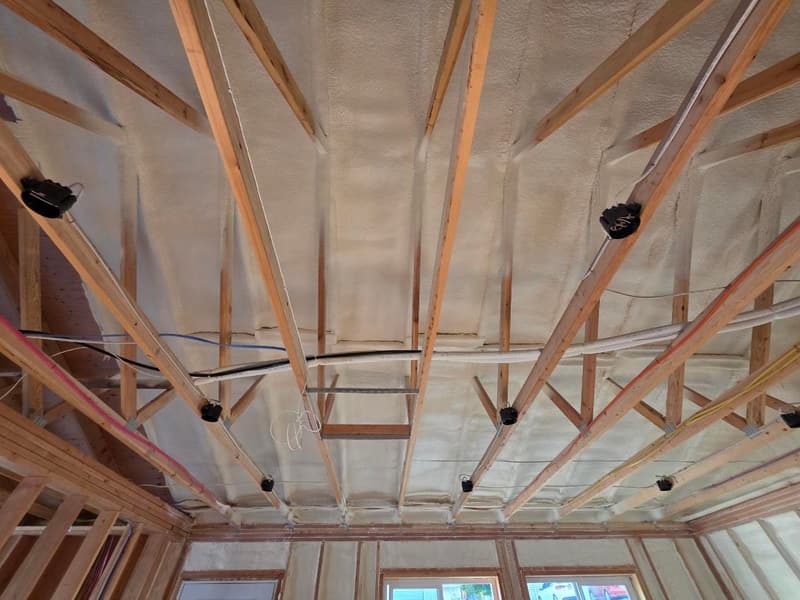 Unfinished ceiling framing with wiring and light boxes, spray foam on the roof deck above and on the walls around the windows