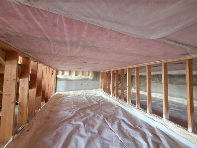 Crawl space with white plastic sheeting over the ground, framed stud walls on both sides and foam along the far foundation wall