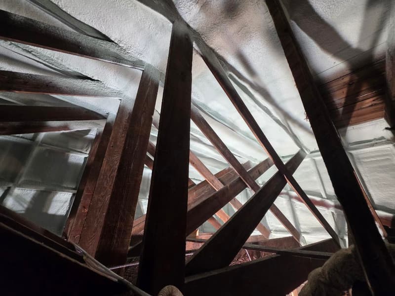 Dark-stained wood trusses with white spray foam on the underside of the roof deck and a spray hose at the edge