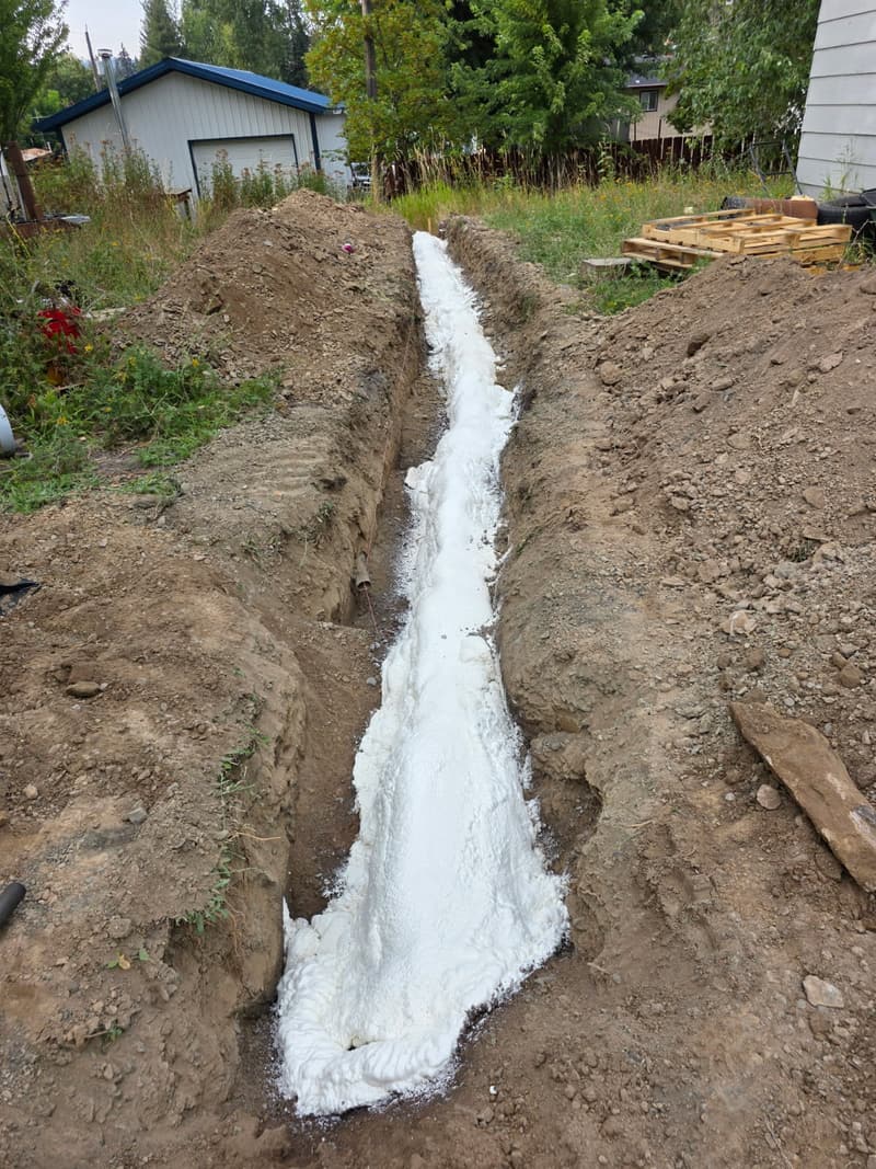 Narrow trench dug through a yard with white spray foam filling its bottom and dirt piled along both sides