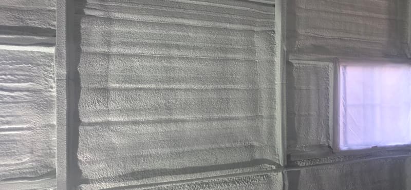 Close view of spray foam built up over a wall's posts and horizontal girts, beside a window masked with plastic