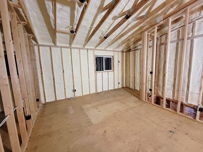 Unfinished room with spray foam filling the wall cavities and the bays between exposed ceiling joists, plywood subfloor below