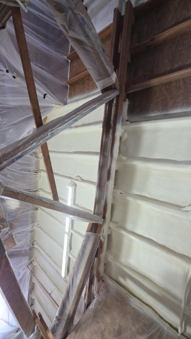 Looking up a corner where spray foam covers a wall between horizontal girts, with plastic sheeting draped over nearby framing