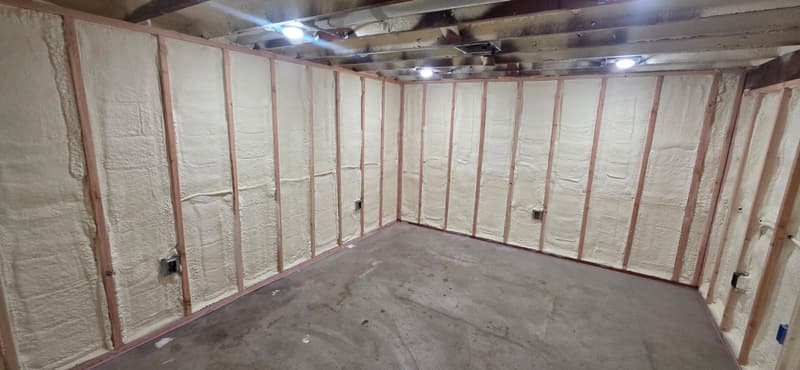 Garage room with spray foam between the wall studs around electrical boxes, open ceiling joists above, concrete floor