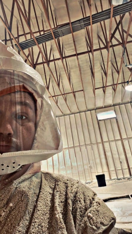 Spray foam installer in a full hood and foam-speckled coveralls inside a building with foamed walls and roof framing
