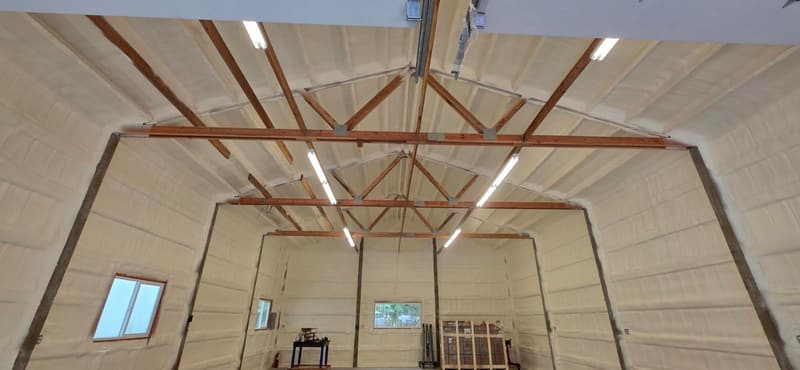 Large building with spray foam on the walls and roof, wood trusses and light fixtures overhead, windows at the far end