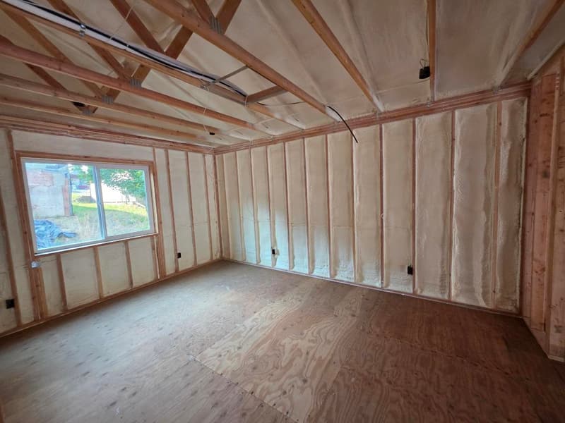 Unfinished room with spray foam between the wall studs, a window, a plywood subfloor and open ceiling framing with wiring