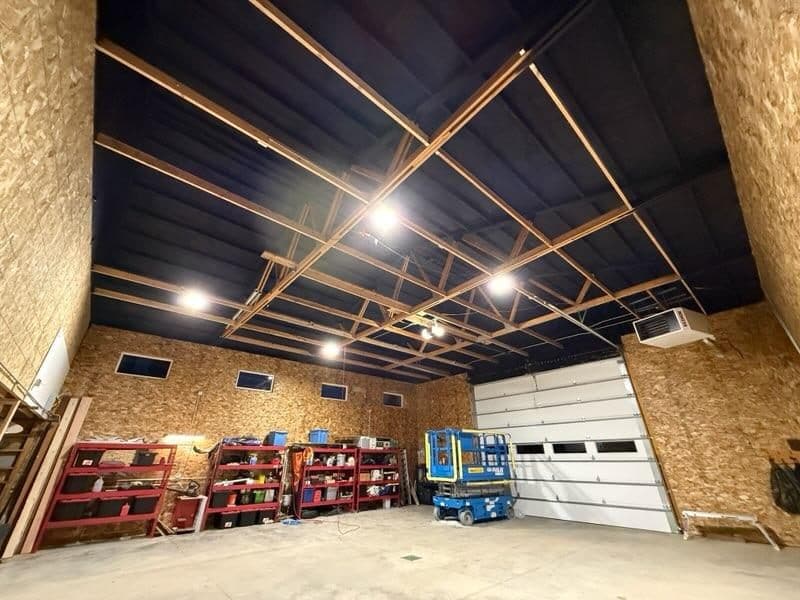 Large shop with OSB-lined walls, a dark ceiling above open wood framing, a white overhead door and a blue scissor lift