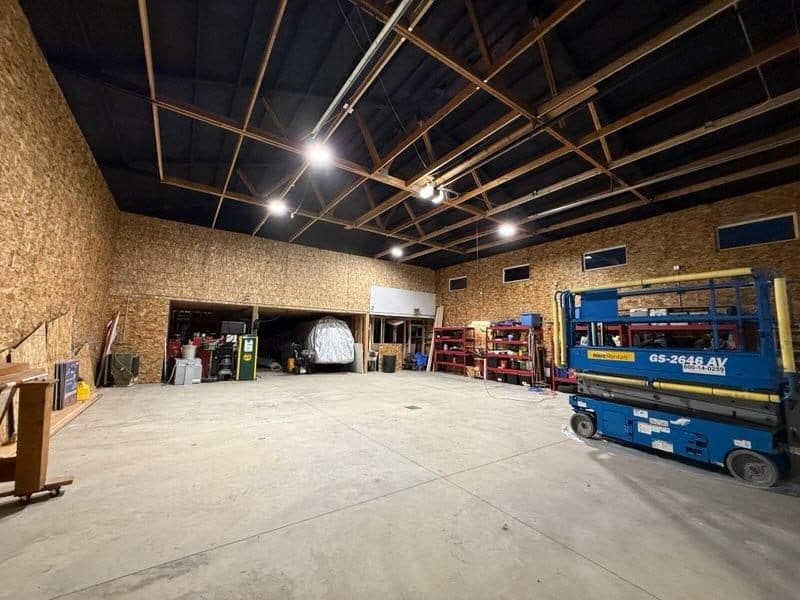 Large shop from another corner: OSB-lined walls, a dark ceiling above open wood framing, open bays and a blue scissor lift