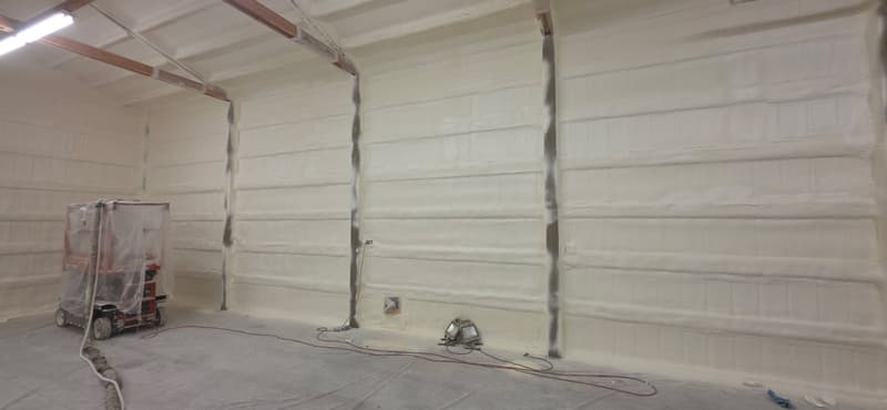Tall building wall freshly sprayed with foam, with a plastic-wrapped spray rig and hoses on the masked floor