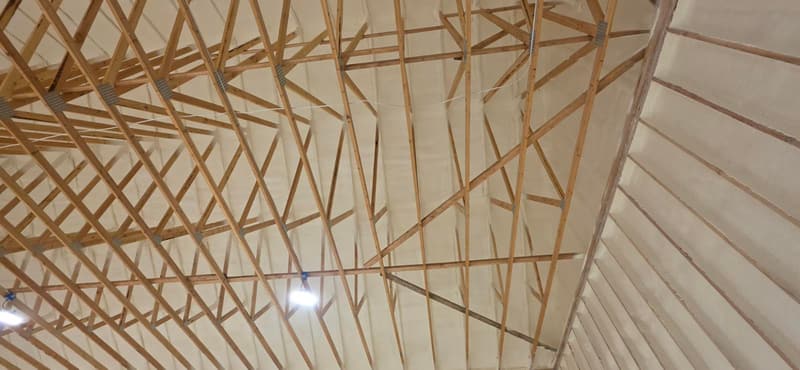 Wide view up into wood roof trusses, with spray foam covering the underside of the roof and the sloped sides