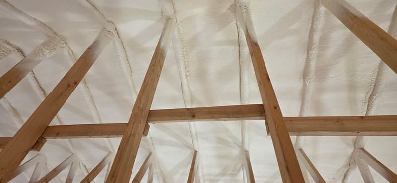 Close view of wood truss members below a roof sprayed with foam, the foam following the framing above