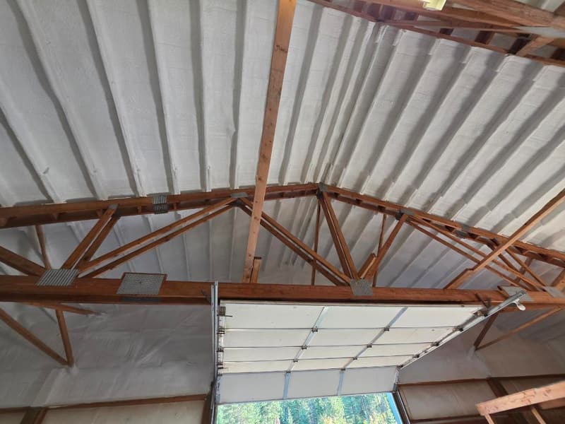 Wood trusses under a roof sprayed with foam that follows the roofing ribs, above a raised overhead door